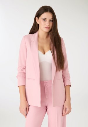 Giovane donna con lunghi capelli castani che indossa un blazer rosa e pantaloni coordinati sopra una maglietta bianca, in piedi su uno sfondo semplice.