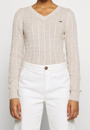Personne portant un pull beige en maille torsadée à col en V et un pantalon blanc taille haute, debout devant un fond uni.