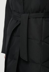 Černá péřová bunda s páskem v pase, zdobená prošitým vzorem a hladkým povrchem. Pásek je uvázán vpředu, čímž zvýrazňuje siluetu.