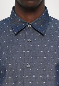 Camisa de botones en denim oscuro con cuello azul, que presenta patrones bordados en blanco de A y X, y un botón gris en el cuello.