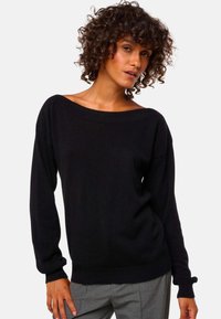 Frau mit lockigem Haar, die einen schwarzen schulterfreien Langarm-Pullover und graue Hosen trägt, steht vor einem einfarbigen Hintergrund.