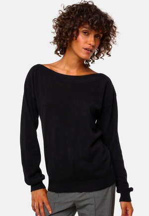 Femme aux cheveux bouclés portant un pull noir à épaules dénudées à manches longues et un pantalon gris, se tenant devant un fond uni.