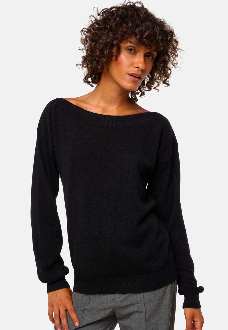 Frau mit lockigem Haar, die einen schwarzen schulterfreien Langarm-Pullover und graue Hosen trägt, steht vor einem einfarbigen Hintergrund.