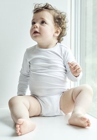 Hvid langærmet skjorte og hvid ble på en siddende tumling. Krøllet hår, lys hud og runde kinder. Glatte teksturer og afslappet holdning.