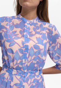 Chemise à manches longues en tissu transparent avec des motifs de feuilles bleues et roses, un col mandarin et un bouton sur le devant. Texture lisse et coupe fluide.