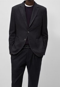 Uomo che indossa un blazer grigio scuro con tasche a toppa, un maglione scuro, una cintura nera e pantaloni neri, in piedi con le mani in tasca.