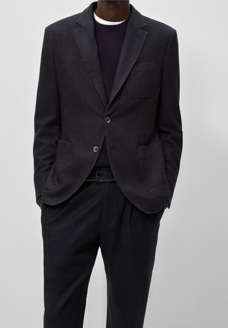 Uomo che indossa un blazer grigio scuro con tasche a toppa, un maglione scuro, una cintura nera e pantaloni neri, in piedi con le mani in tasca.