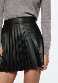 Jupe en cuir noir plissée avec une taille lisse, présentant des plis verticaux aigus et une texture lisse et brillante.
