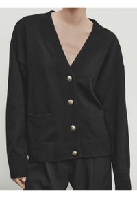 Cardigan noir à col en V avec boutons dorés, manches longues et deux poches avant, porté avec un pantalon noir.