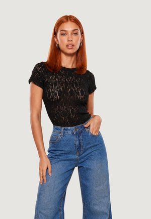 Femme aux cheveux roux lisses portant un haut noir en dentelle à manches courtes et un jean bleu taille haute à jambes larges, debout avec une main dans la poche.