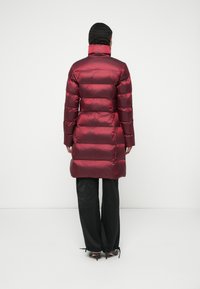 Vínový péřový kabát s horizontálním prošíváním, vysokým límcem a dlouhými rukávy. Nosit s černými kalhotami a podpatky. Hladký, lesklý povrch.