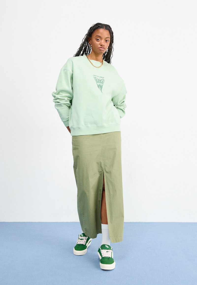 Sweatshirt vert clair avec un graphique de papillon, associé à une jupe olive mi-longue avec une fente à l'avant. Chaussettes blanches et baskets vertes.