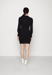 Femme aux cheveux noirs attachés en queue de cheval basse, portant une mini-robe noire à manches longues côtelée et des bottines blanches, debout devant un fond uni.