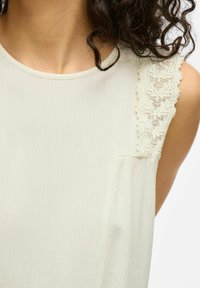 Blusa sin mangas de color crema claro con tejido texturizado, detalles de encaje en el hombro y un pequeño bolsillo delantero. Apariencia suave y lisa.