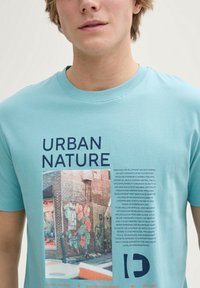 Šviesiai mėlynas medvilninis marškinėliai su grafiniu spaudiniu, kuriame yra tekstas "URBAN NATURE" ir spalvota freskos nuotrauka ant mūrinės sienos.