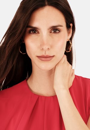 Ung kvinde med langt brunt hår iført gyldne bølgede hoops-ørestikker og en rød bluse, der blidt rører ved sin hals, med et neutralt udtryk.