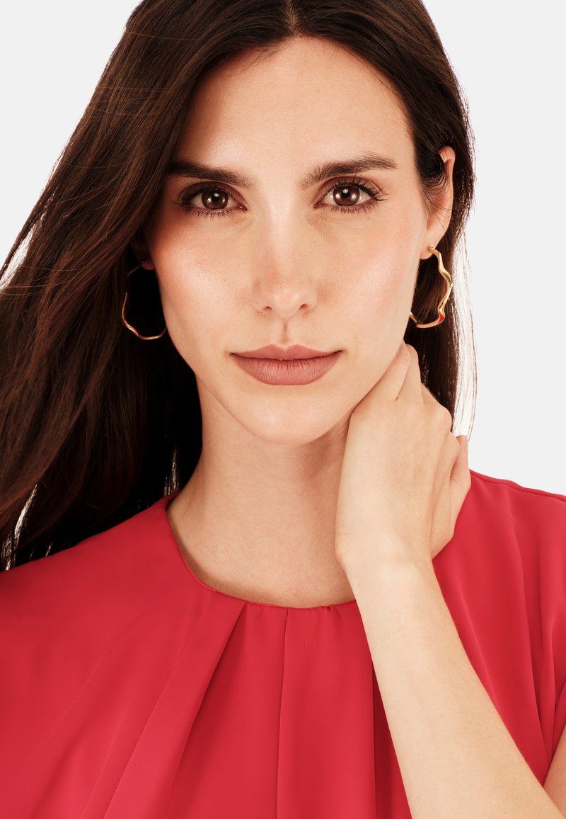 Ung kvinde med langt brunt hår iført gyldne bølgede hoops-ørestikker og en rød bluse, der blidt rører ved sin hals, med et neutralt udtryk.