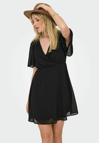 Robe noire avec un design enroulé, manches courtes volantées, tissu doux, et une jupe évasée qui tombe au-dessus du genou. Accentuée d'un chapeau brun.
