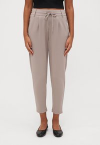 Lichtbeige taps toelopende broek met een elastische trekkoord taille; heeft plooien en een soepele textuur, gecombineerd met zwarte platte schoenen.