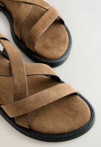 Sandales en daim marron avec des brides croisées, semelles en caoutchouc noir et une texture lisse. Design minimaliste sans quincaillerie visible.