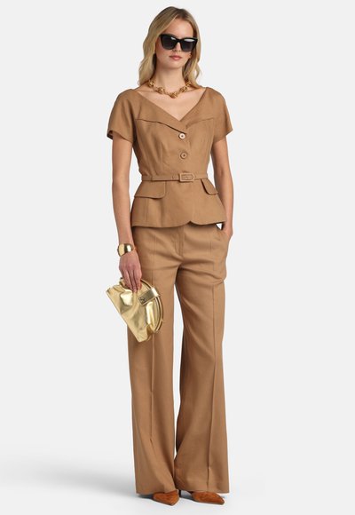 Woman wearing tan belted short-sleeve top and matching wide-leg pants, holding gold clutch, with black sunglasses and gold jewelry.