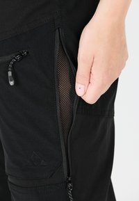 Musta cargo-housut sisältävät vetoketjullisen taskun, jossa on verkkomateriaali, ja niissä on sileä pinta sekä erottuva tuotemerkin logo painettuna kankaaseen.
