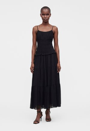 Femme portant une robe longue noire à volants sans manches avec de fines bretelles, debout droite sur un fond clair uni, portant des talons noirs à lanières.