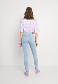 Camiseta de tie-dye en color púrpura claro con mangas cortas, combinada con jeans ajustados azul claro que cuentan con dos bolsillos traseros y una etiqueta de cuero marrón. Zapatillas rosas.