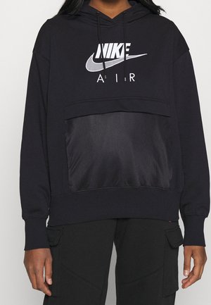 Persoon draagt een zwarte Nike Air-hoodie met gaas voorvak en bijpassende zwarte cargobroek, zichtbaar van schouders tot heupen.