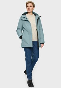 Frau mit einer hellblauen Winterjacke mit Kapuze, beigem Pullover, blauen Jeans und schwarzen Stiefeln, die mit den Händen in den Jackentaschen steht.