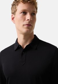 Młody mężczyzna z krótkimi blond włosami, ubrany w czarną koszulę z kołnierzykiem, patrzący lekko na bok na tle jasnego, jednolitego tła.