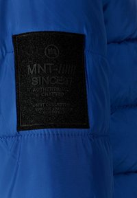 Mėlynas pūkinis džemperis su juodu stačiakampiu etikete, ant kurios įspaustas tekstas. Medžiaga yra lygi ir šiek tiek atspindinti, matomos siūlės.