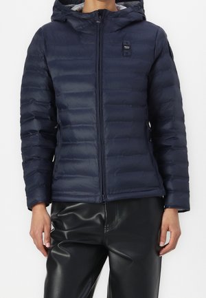 Personne portant une veste matelassée à capuche bleu marine avec fermeture éclair et un pantalon en cuir noir, debout devant un fond uni.