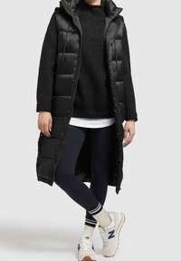 Czarna kamizelka puchowa z kapturem, pikowany wzór, zamek z przodu, noszona na czarnym swetrze, granatowych legginsach i białych sneakersach z granatowymi akcentami.