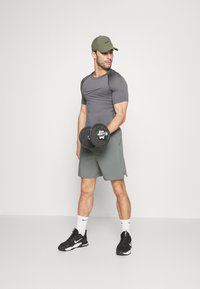 Camisa atlética cinza justa com mangas curtas, calções verdes, ténis pretos e um boné verde. Segurando um haltere preto com uma mão.