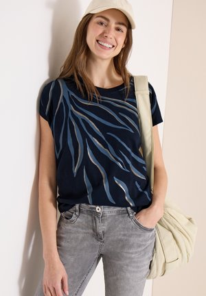 Donna sorridente che indossa un cappellino beige, una camicia blu navy a fantasia, jeans grigi e porta una borsa a tracolla color crema.