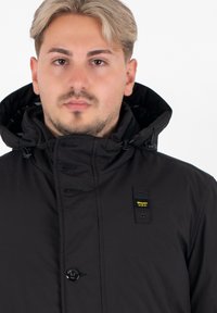 Zwarte hoodie-jack met een knoop- en drukknoopsluiting, voorzien van een zichtbaar merkembleem op de borst en aantrekkoorden in de capuchon.
