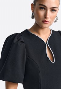 Schwarzes Kleid mit Puffärmeln, die Stoffstruktur ist sichtbar; es hat einen Schlüsselloch-Ausschnitt, der mit einer Reihe klarer Verzierungen geschmückt ist.