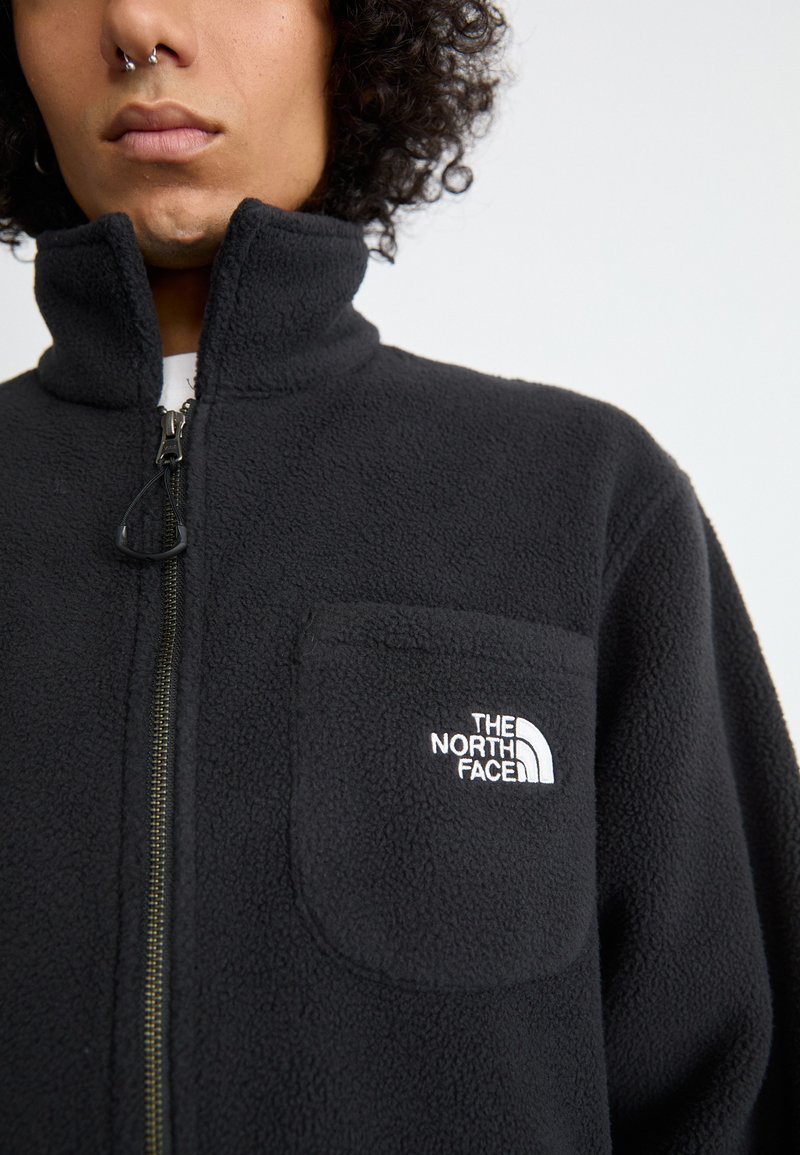 Veste en polaire noire avec un col haut, fermeture éclair à l'avant et une poche poitrine arborant un logo blanc. Texture douce et design décontracté.