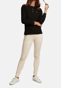 Černé polokošile s dlouhým rukávem a límcem, bílé knoflíky, v kombinaci s béžovými slim džíny a bílými teniskami, stojící postava.