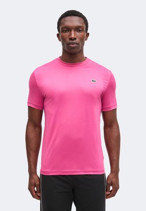 Mann trägt ein leuchtend pinkes, enganliegendes Kurzarmhemd mit einem kleinen grünen Krokodil-Logo auf der Brust, kombiniert mit schwarzen Hosen.