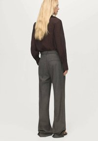 Camicia lunga a maniche lunghe di un marrone scuro e dalla texture morbida, abbinata a pantaloni grigi a vita alta e a gamba larga con tasche sul retro e cuciture rifinite.