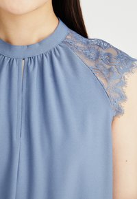Top azzurro chiaro con scollatura alta e maniche in pizzo trasparente, caratterizzato da delicati motivi floreali e un dettaglio a occhio dritto nella parte anteriore.