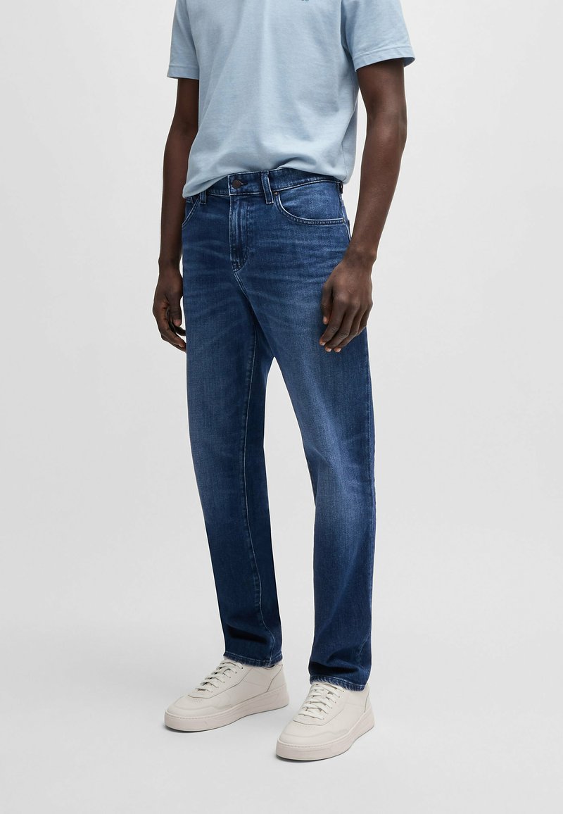 Jeans blu scuro con taglio dritto, caratterizzati da un classico design a cinque tasche e una sfumatura leggera. Abbinati a sneakers bianche.