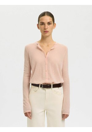 Femme portant un cardigan rose clair boutonné, rentré dans un pantalon crème taille haute avec une ceinture foncée, debout devant un fond uni.