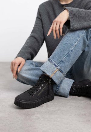Zapatillas altas negras con una suela gruesa y estriada, parte delantera con cordones y detalle de cremallera lateral. Llevadas con jeans azul claro doblados en el dobladillo.