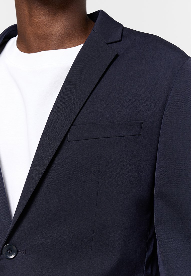 Blaser bleu marine avec une texture lisse, fermeture à un bouton et une poche poitrine ; porté sur un T-shirt blanc, avec un revers cranté.
