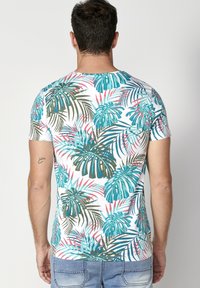 Camiseta de manga corta en blanco con un patrón de hojas tropicales multicolores en teal, verde y rosa; diseño ajustado, tela suave, cuello redondo.