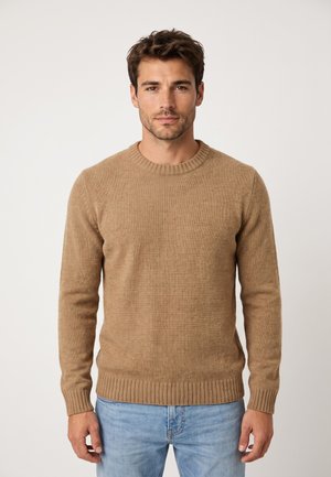 Hombre con el pelo corto y castaño, vestido con un suéter de punto beige y vaqueros azul claro, de pie frente a un fondo claro y sencillo.
