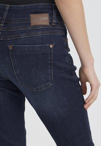 Vista trasera de una persona que lleva vaqueros de mezclilla azul oscuro con parche de cuero de la marca y costuras visibles en el bolsillo trasero.